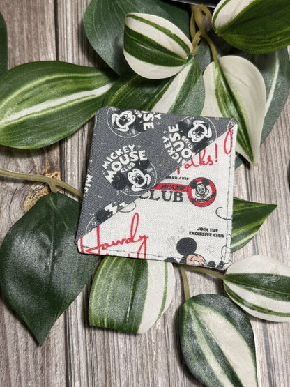 A square fabric book mark with a Mickey Mouse Club graphic print, styled on a rustic wood background with green and white variegated plant leaves.