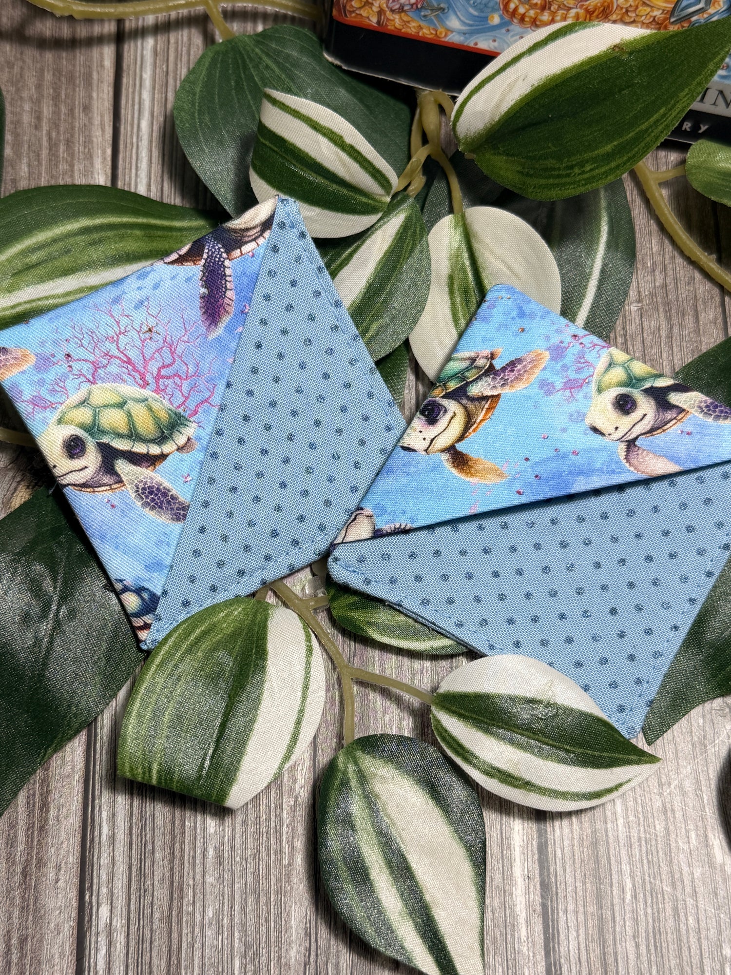 Two book marks with a blue and green sea turtle pattern, displayed on a rustic wood surface surrounded by green and white variegated houseplant leaves.
