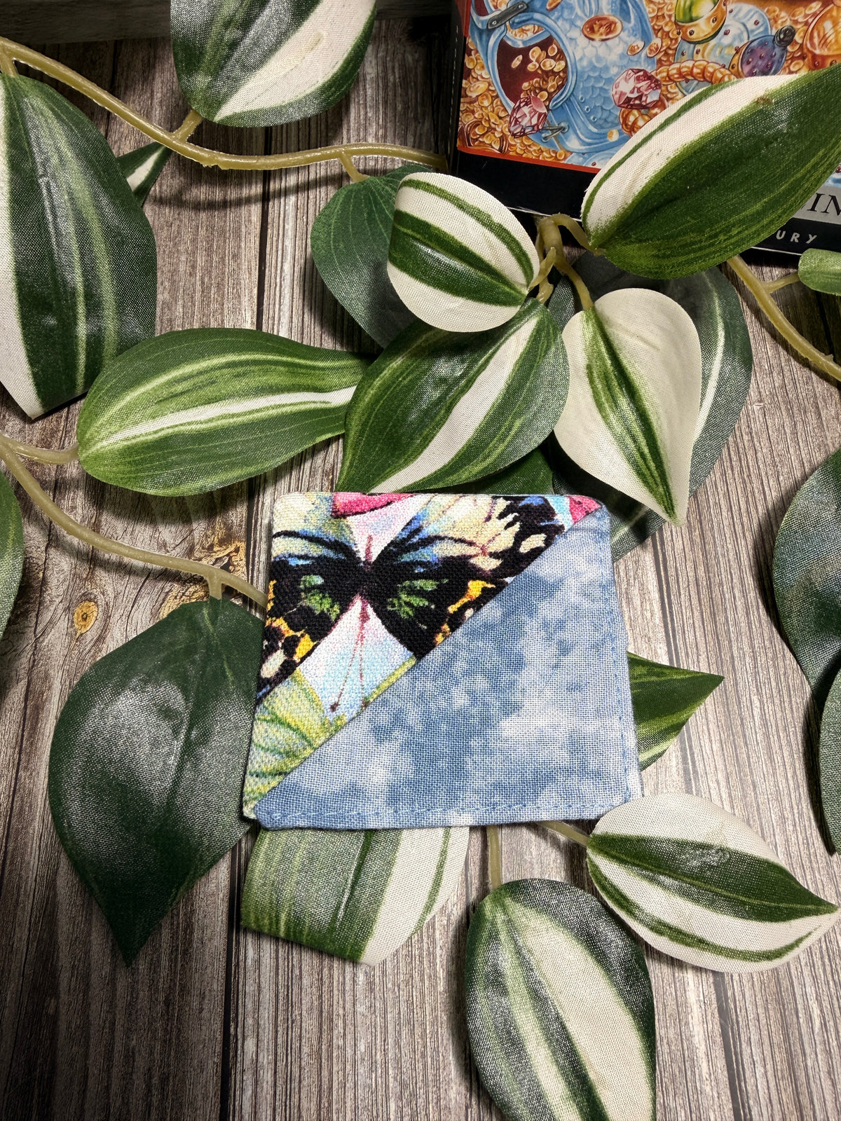 A square fabric book mark with a diagonal split design, featuring a butterfly print on one side and a light blue marbled pattern on the other, styled on a wooden surface with green variegated leaves.