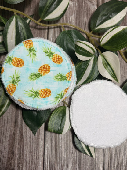 Two reusable cloth make up remover wipes with a light blue pineapple print and a white plush side, displayed on a rustic wood background with green variegated leaves.