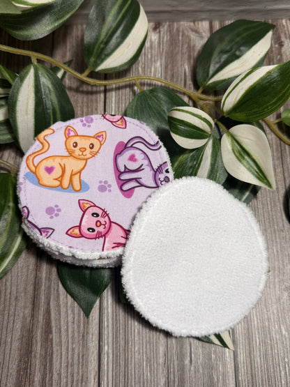 TwoRound fabric face cleanser and make up remover pads with a fuzzy white backing, one showing a light purple cover printed with cartoon orange, pink, and purple cats, and the other showing the plain white fuzzy side, displayed on a wooden surface with green variegated leaves.