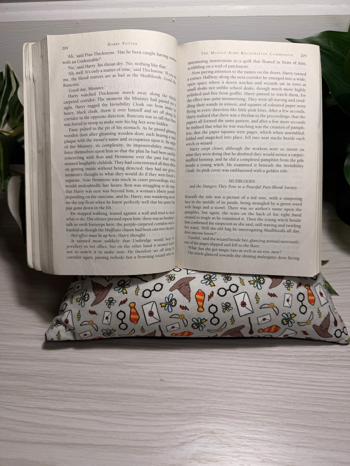 An open book lying on a patterned fabric cushion on a light-colored wooden surface, with a green leaf visible in the background.