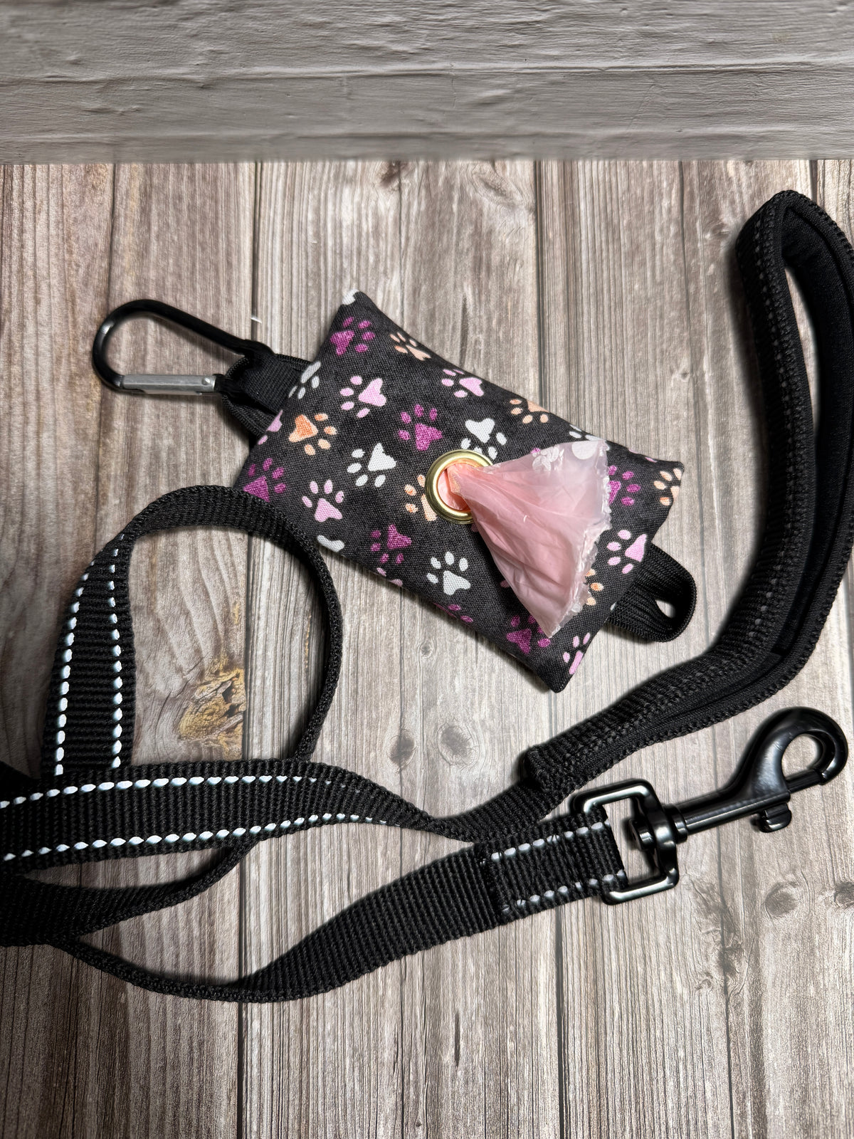A black dog leash with white reflective stitching and a matching black clip, shown with a small patterned poop bag holder containing pink dog poop bags, laid flat on a gray wood plank background.