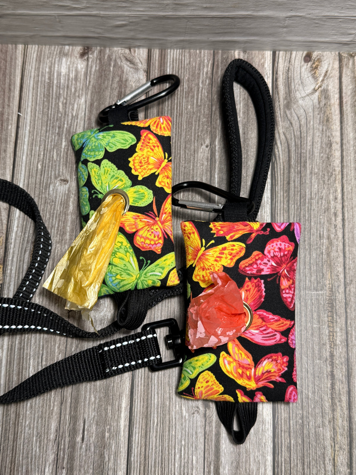 Two small black fabric pet waste bags with colorful butterfly and floral prints, shown on a gray wood floor. Each bag has a black carabiner clip and a black strap, with one bag also showing a gold-colored plastic pull tab.