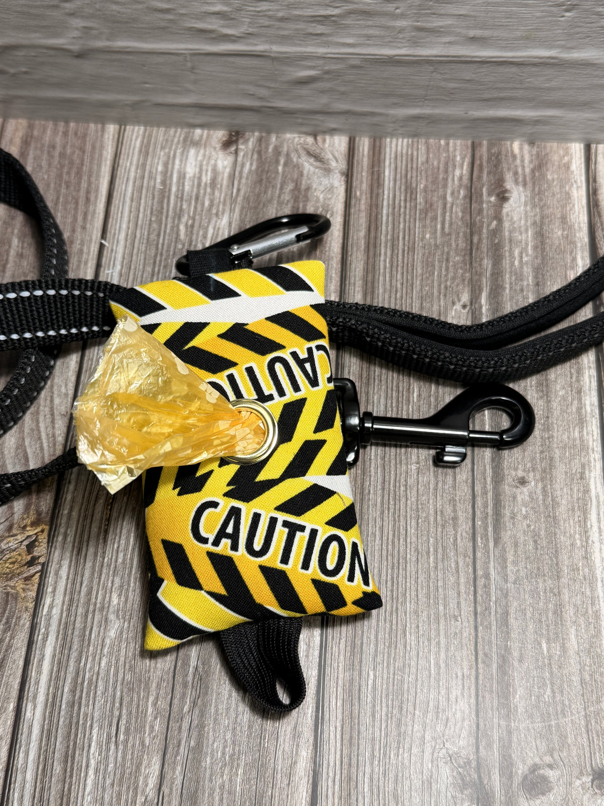 A yellow and black caution tape dog poop bag with 'CAUTION' printed on it, attached to a black strap and shown on a gray wood-plank floor with a piece of trash inserted.