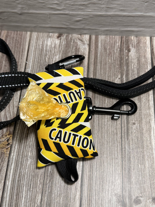 A yellow and black caution tape dog poop bag with 'CAUTION' printed on it, attached to a black strap and shown on a gray wood-plank floor with a piece of trash inserted.