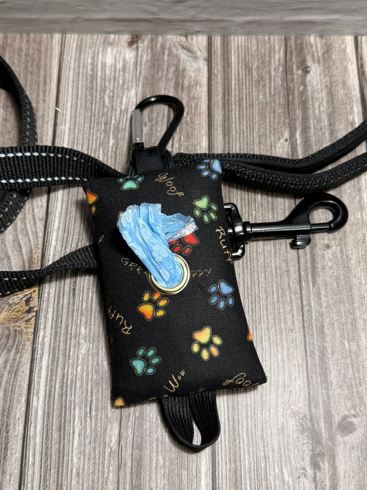 A black fabric dog leash poop bag pouch with colorful paw print patterns, a blue leash collar, and a black carabiner, shown with a black leash on a gray wood plank background.
