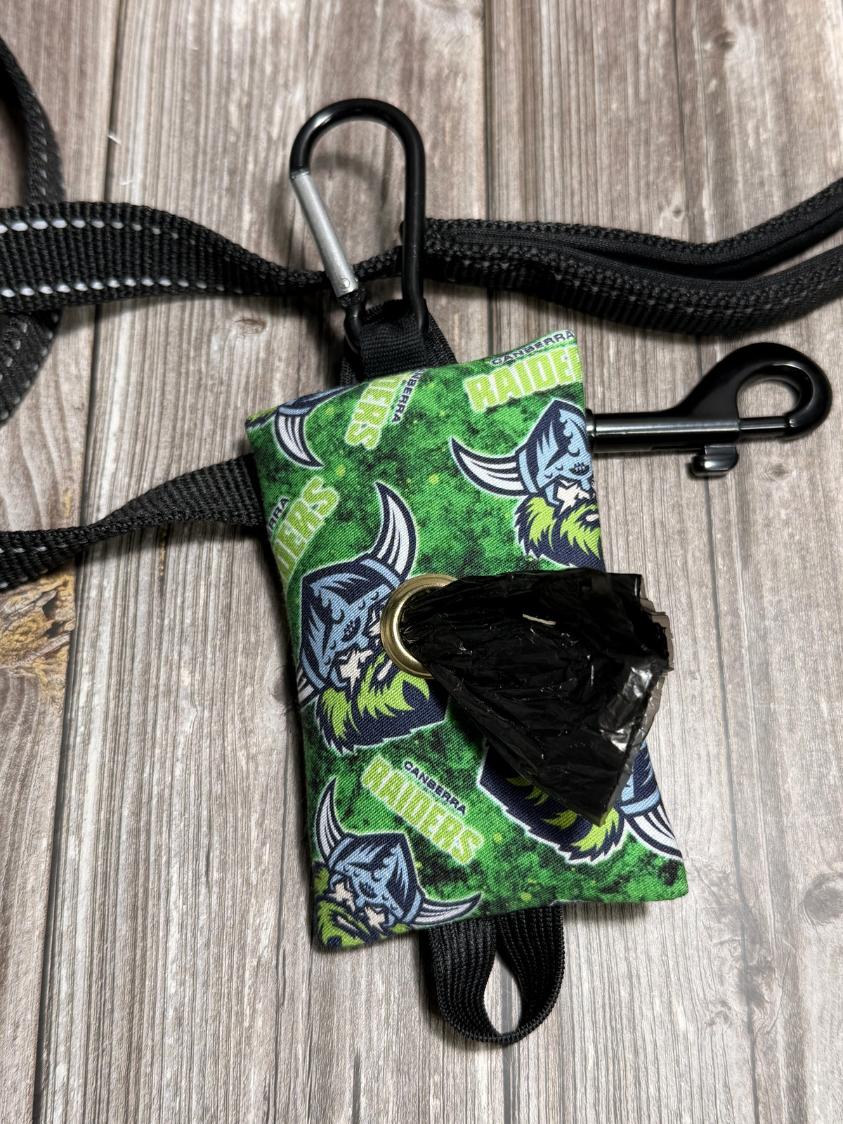 A dog leash and leash bag set with a green and black patterned fabric bag holding a black plastic bag, attached to black nylon straps and a black carabiner, on a gray wood plank background.