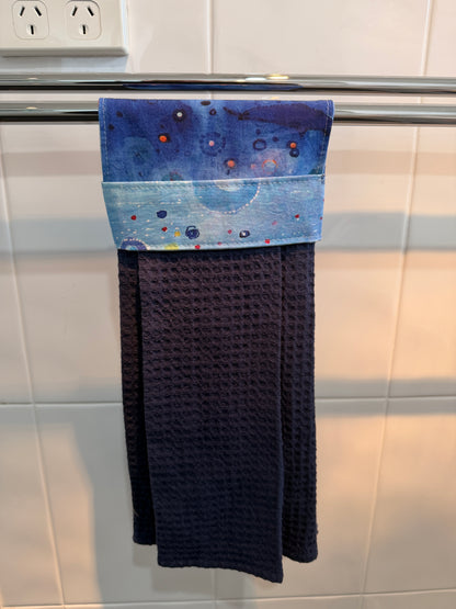 A dark navy blue waffle-weave kitchen towel hanging from a metal rack on a white tiled wall. The top of the towel features a decorative band with a blue and white ocean pattern.