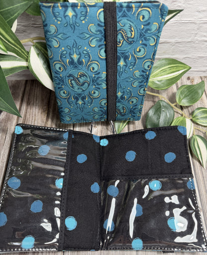 a needle book with a teal and gold dragon design sitting against a brick wall, with an open needle book showing clear pockets and a black with teal spots design with on a wooden surface.