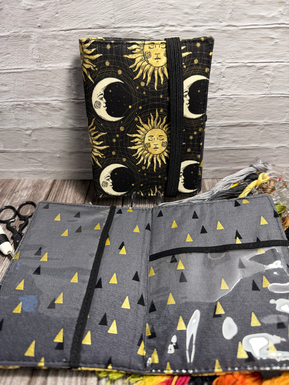 a needle book with a sun and moon patterned design sitting against a brick wall, with an open needle book showing clear pockets and a grey with gold and black triangle design with cross stitching supplies underneath.