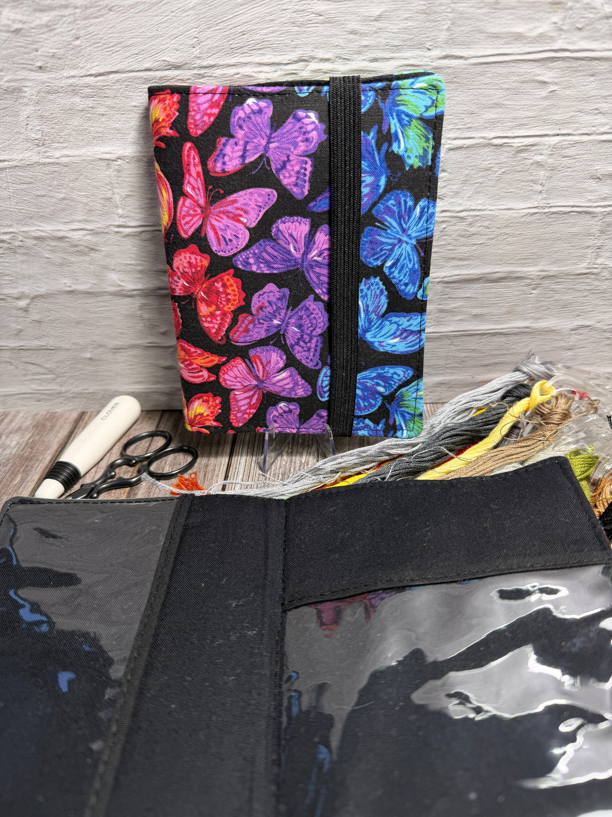 a needle book with a colourful butterfly design sitting against a brick wall, with an open needle book showing clear pockets and a black fabric background with cross stitching supplies underneath.