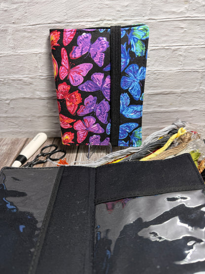 a needle book with a colourful butterfly design sitting against a brick wall, with an open needle book showing clear pockets and a black fabric background with cross stitching supplies underneath.