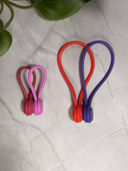 Colorful silicone needle minders in a small size and large size on a textured surface with a plant in the corner.