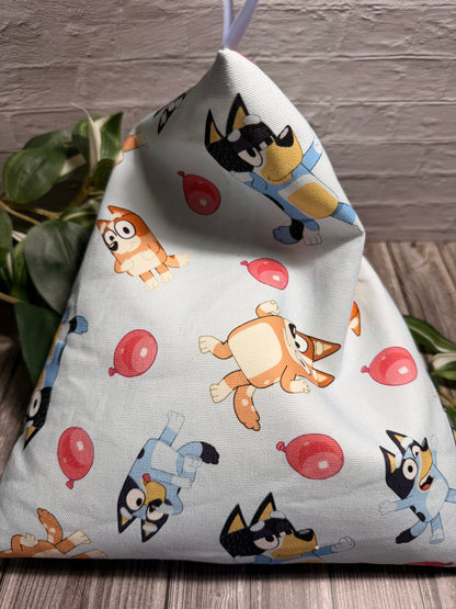 A triangular fabric lap pillow with a light blue-gray background and a colorful cartoon bluey and bingo dog print, shown standing upright on a wooden surface with green leaves and a brick wall in the background.