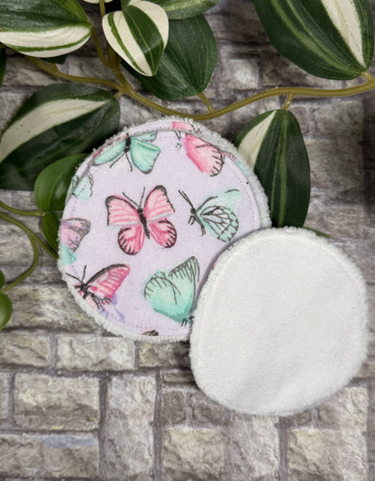 Two Round fabric face cleanser and make up remover pads with butterfly design on a stone surface with green leaves.