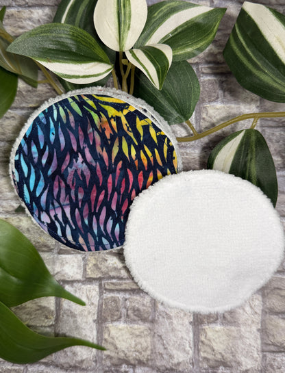 two Round fabric face cleanser and make up remover pads, one with a colorful pattern and the other white, on a stone surface with green leaves.