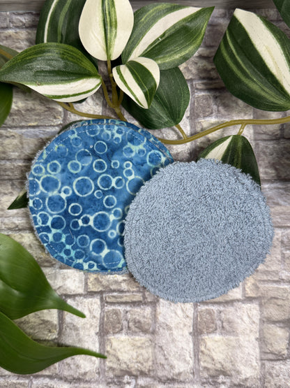 Two Round fabric face cleanser and make up remover pads with blue bubble pattern on a stone surface with green leaves.