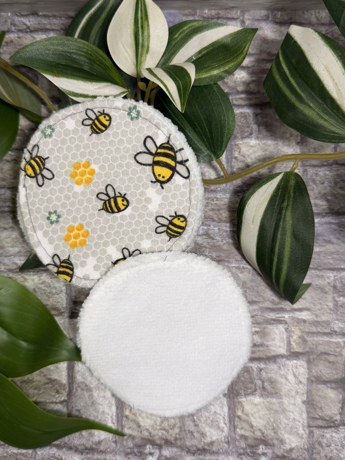 Round fabric face cleanser and make up remover pads with bee designs on a stone surface with green leaves.