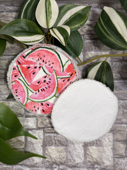 Round fabric face cleanser and make up remover pads with watermelon design on a stone surface with green leaves.