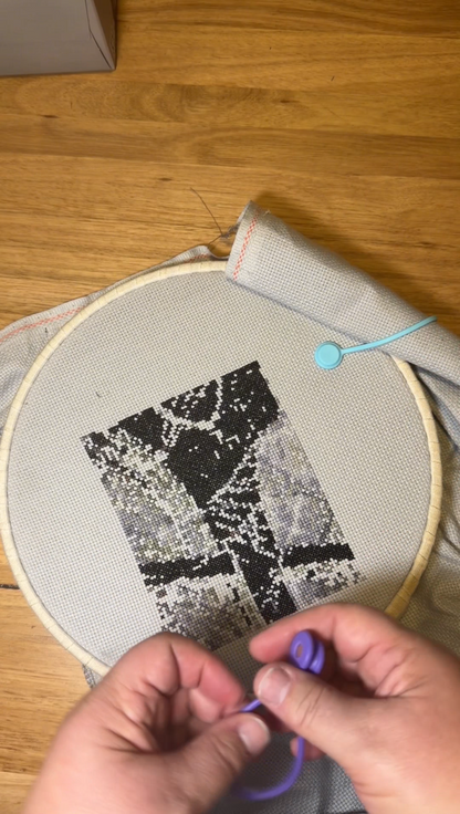 Person working using a silicone needle minder on a cross-stitch project with an embroidery hoop and pattern on a wooden surface.