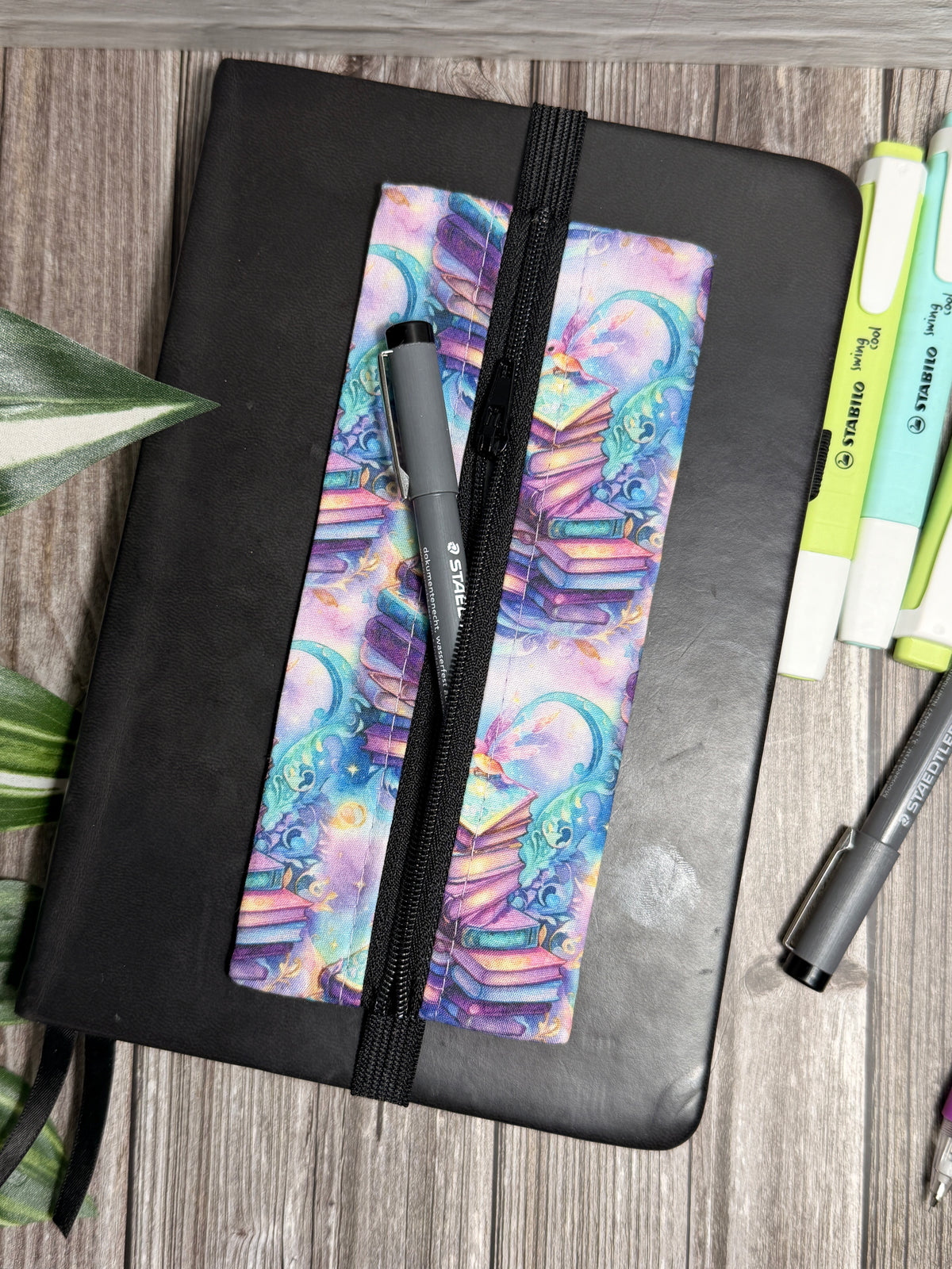 Pencil case with small books which are pinks, purples and teals which is held onto a book with elastic on a wooden surface with highlighters 