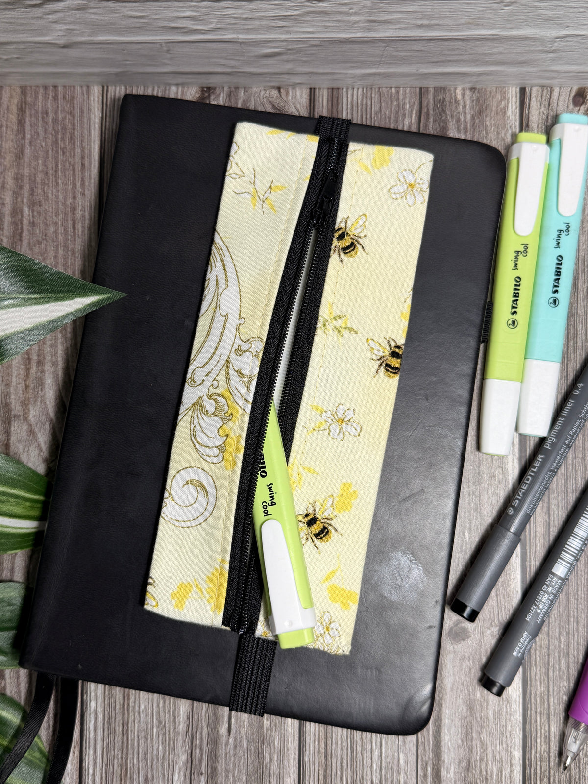 Pencil case with small bees and a filagree and flower design which is held onto a book with elastic on a wooden surface with highlighters 