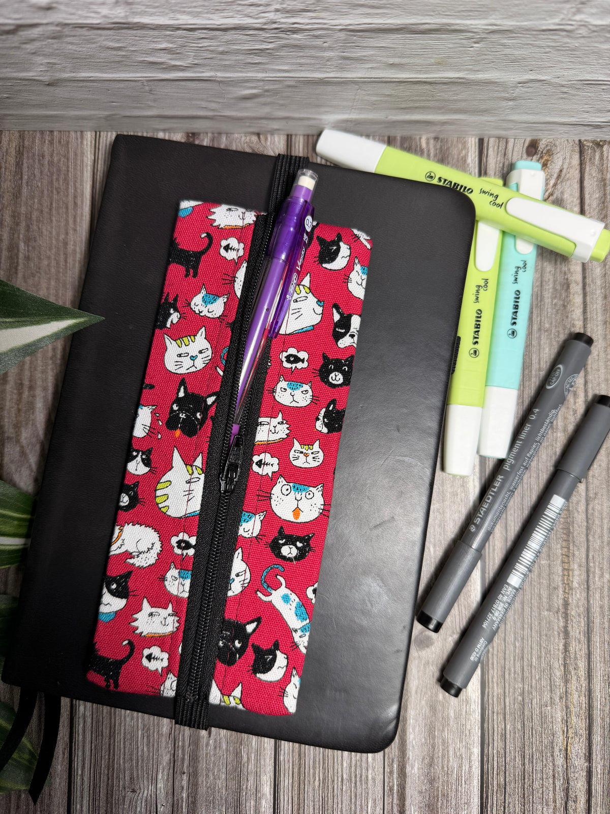 Pencil case with small cats on a dark pink background which is held onto a book with elastic on a wooden surface with highlighters 