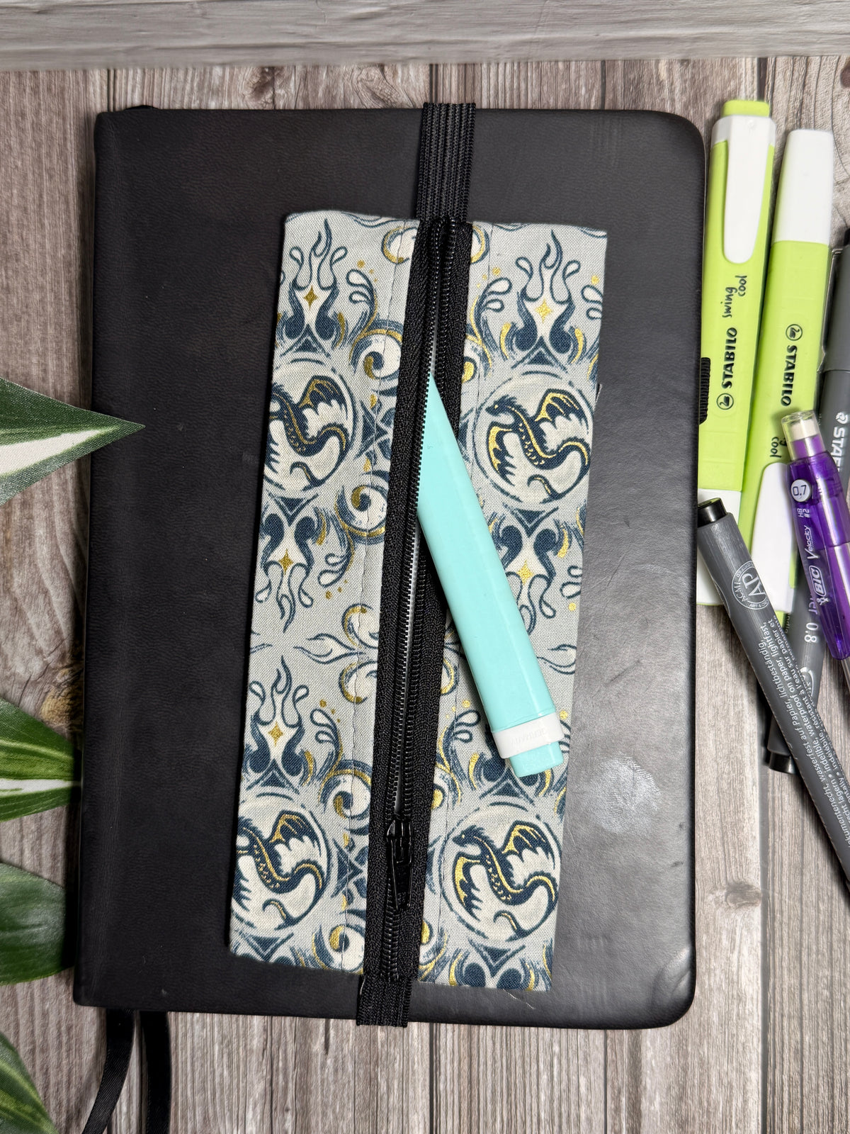 Pencil case with small dragons on a grey and teal background which is held onto a book with elastic on a wooden surface with highlighters