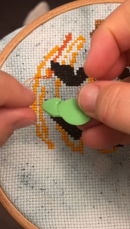 Cross-stitching process with a long looped needle threader held over a partially completed design so help with thread chicken.