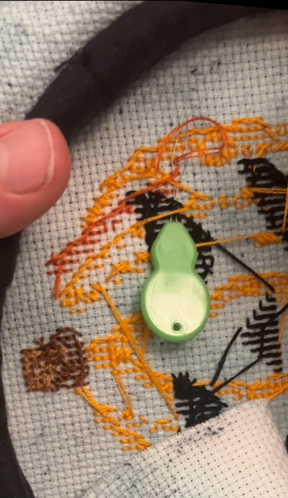 Close-up of a hand holding a cross stitch hoop using a needle threader to tuck short floss tails over an embroidered design.